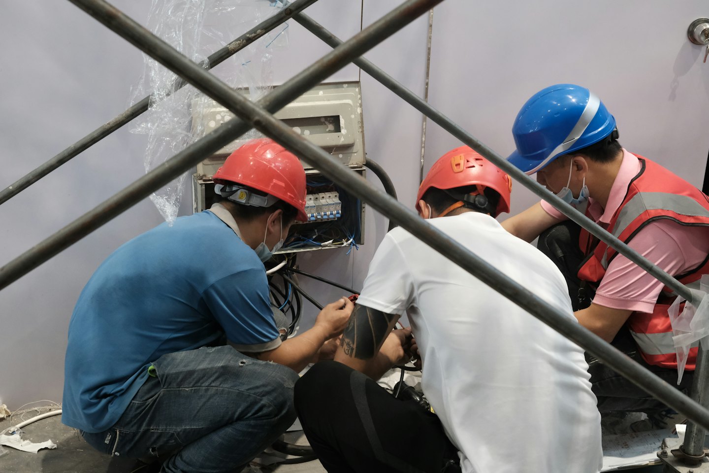 Men in hard hats connect wires to an electrical panel indoors.